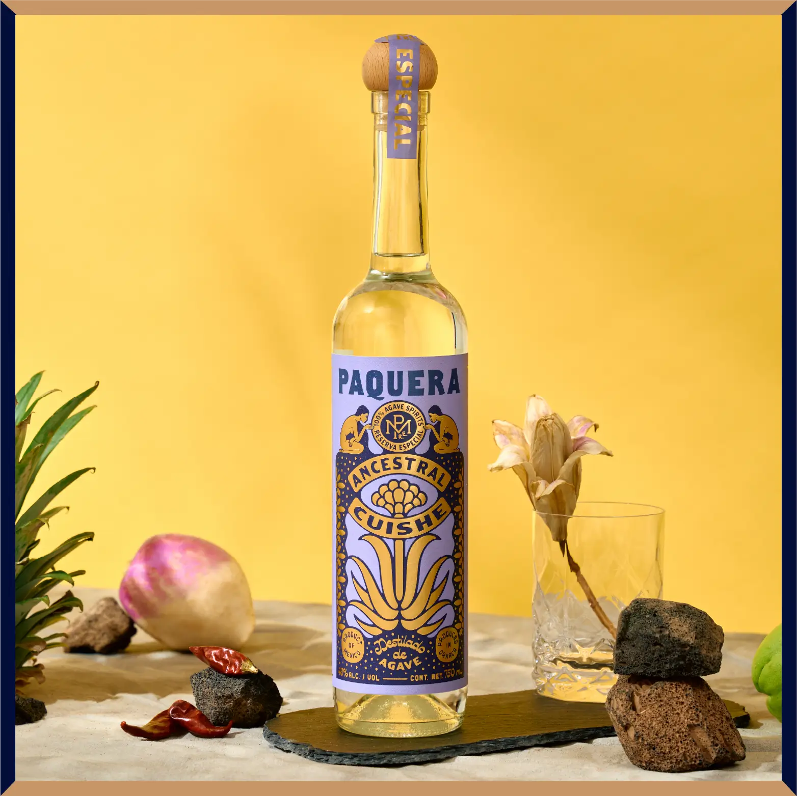 Paquera Mezcal Ancestral Cuishe bottle with purple and gold label, placed on a neutral surface with rocks, pineapple top, dried chili, and tropical produce in front of a yellow background