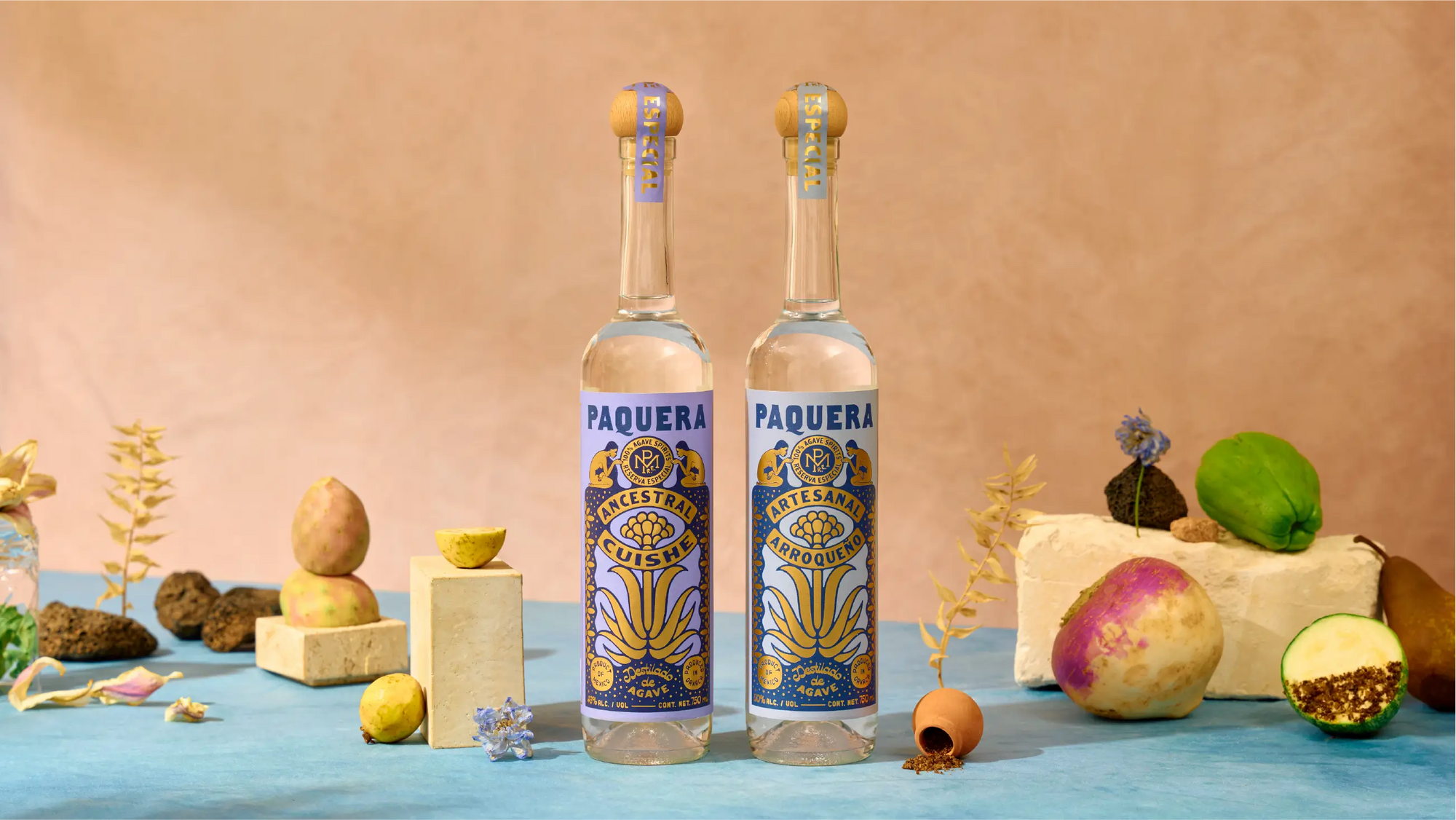 Two Paquera Mezcal bottles, Ancestral Cuishe and Artesanal Arroqueño, displayed with fruits, roots, flowers, and volcanic rock elements on a blue surface and pink backdrop