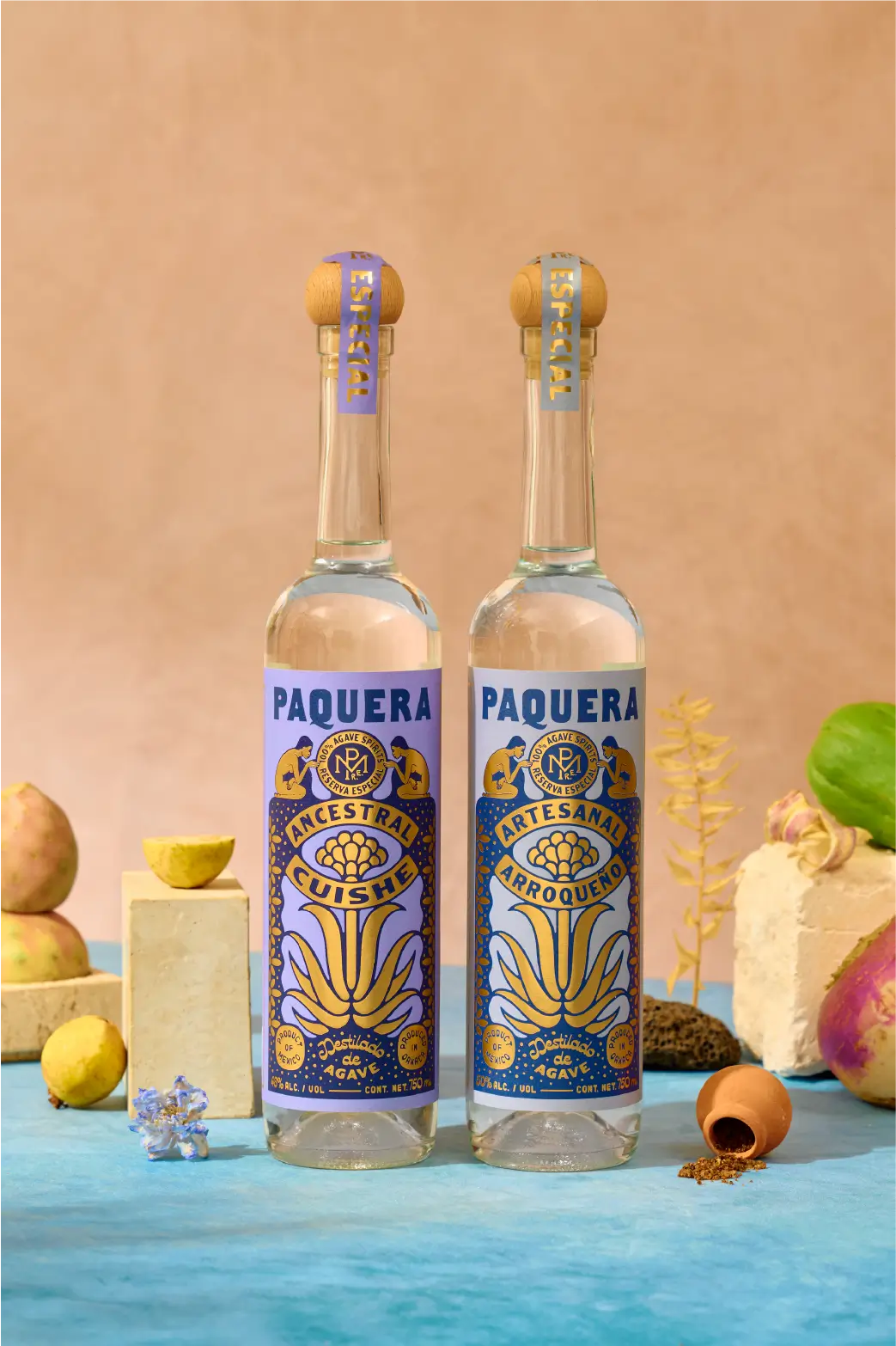 Two Paquera Mezcal bottles, Ancestral Cuishe and Artesanal Arroqueño, displayed with fruits, roots, flowers, and volcanic rock elements on a blue surface and pink backdrop