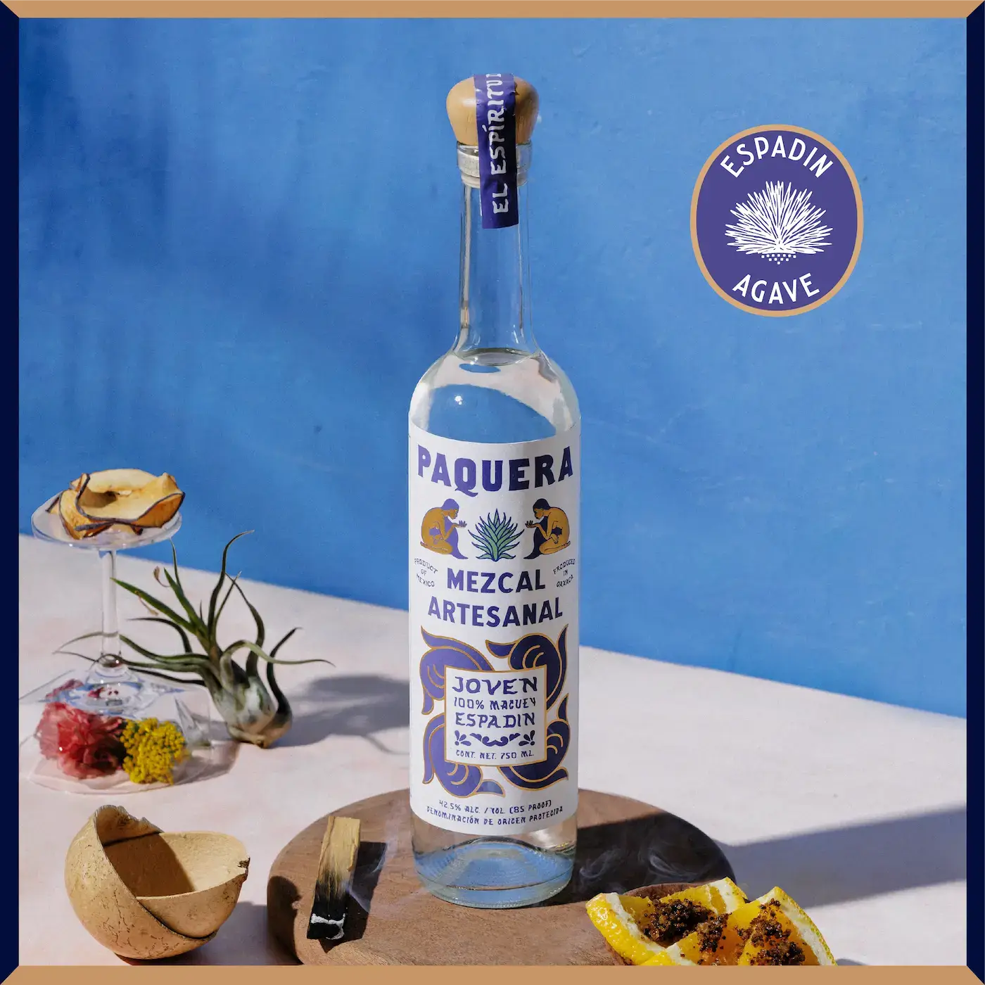 Paquera Mezcal Artesanal Espadín bottle against a blue background, styled with smoked wood, citrus, dried fruit, and botanicals.
