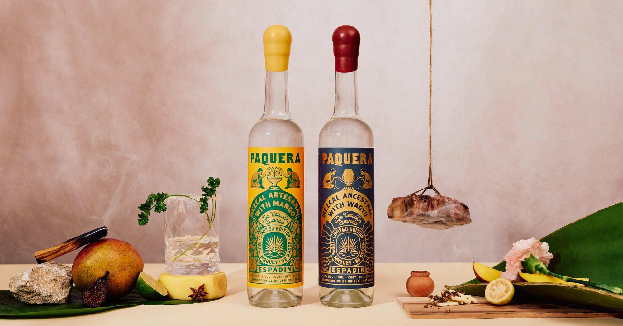 Two bottles of Paquera Mezcal stand side by side, one with a yellow cap and label featuring mango, and the other with a red cap and dark label featuring Wagyu. The labels display intricate illustrations of traditional mezcal-making. Surrounding the bottles are fresh ingredients including mango, lime, and spices, along with a glass of mezcal, a smoking incense stick, and a hanging piece of meat, all set against a warm, neutral-toned background.