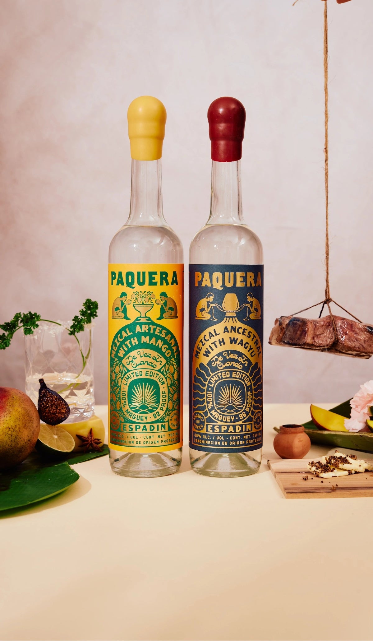 Two bottles of Paquera Mezcal stand side by side, one with a yellow cap and label featuring mango, and the other with a red cap and dark label featuring Wagyu. The labels display intricate illustrations of traditional mezcal-making. Surrounding the bottles are fresh ingredients including mango, lime, and spices, along with a glass of mezcal, and a hanging piece of cured meat, all set against a warm, neutral-toned background.